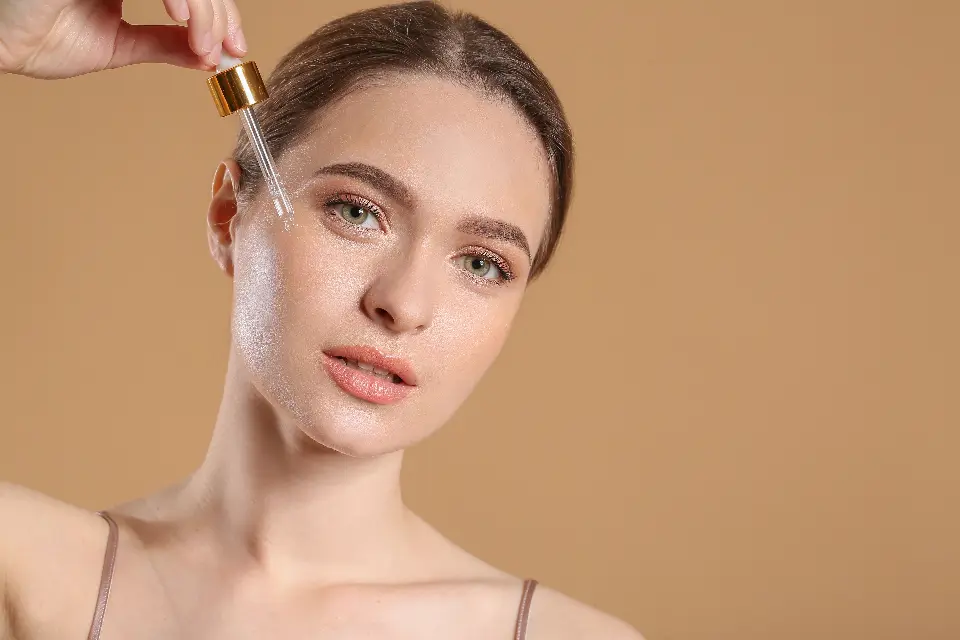 Mulher de pele bem cuidada aplicando sérum facial com conta-gotas, em fundo bege