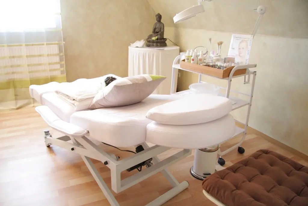 Sala de atendimento com maca branca, almofadas e carrinho com produtos de estética, iluminada por luz natural