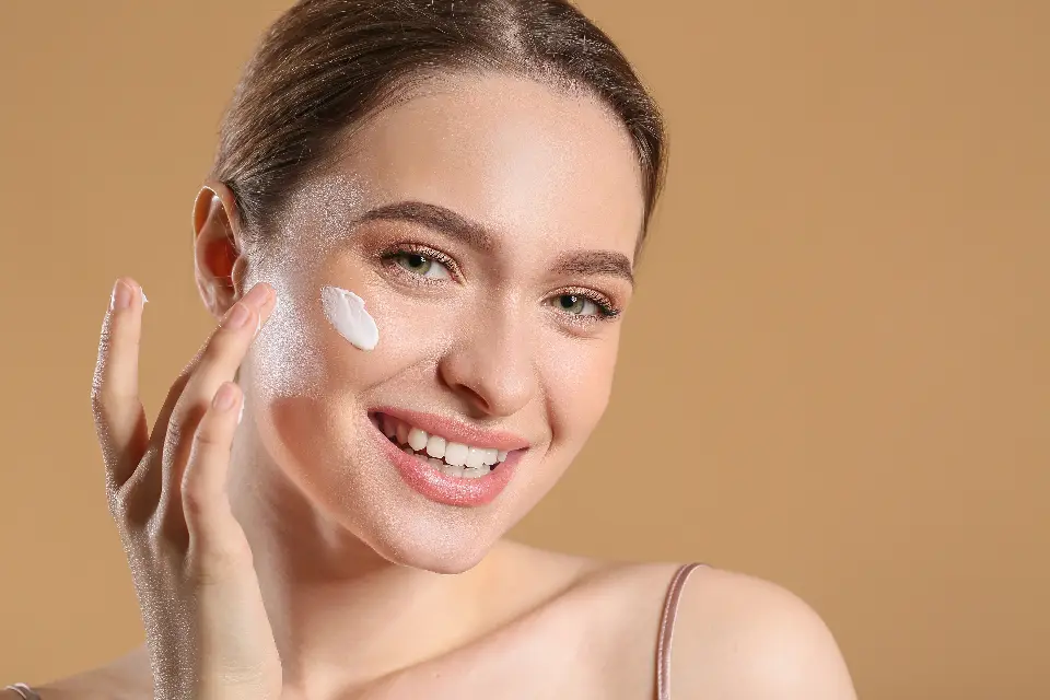 Mulher sorrindo enquanto aplica creme hidratante no rosto, sobre fundo bege
