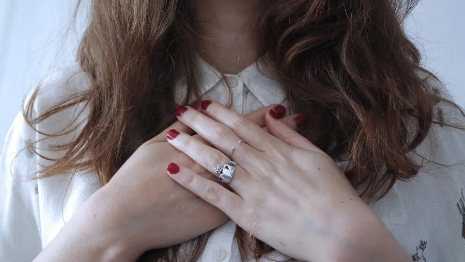 Mãos cruzadas sobre blusa branca, com unhas curtas vermelhas e anéis prateados