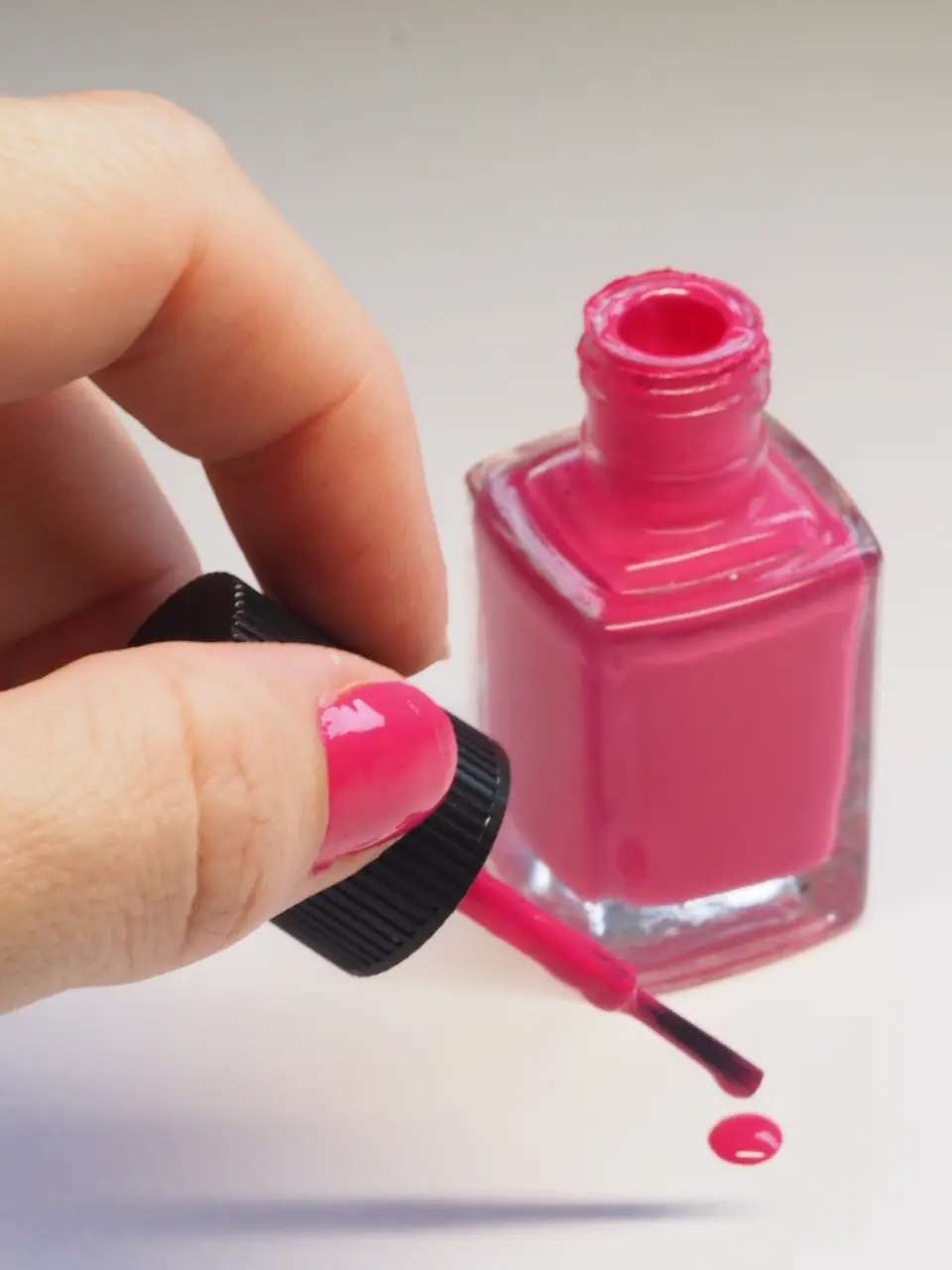 Mão com unha pink brilhante segurando o pincel de um esmalte rosa, com o vidro aberto ao fundo